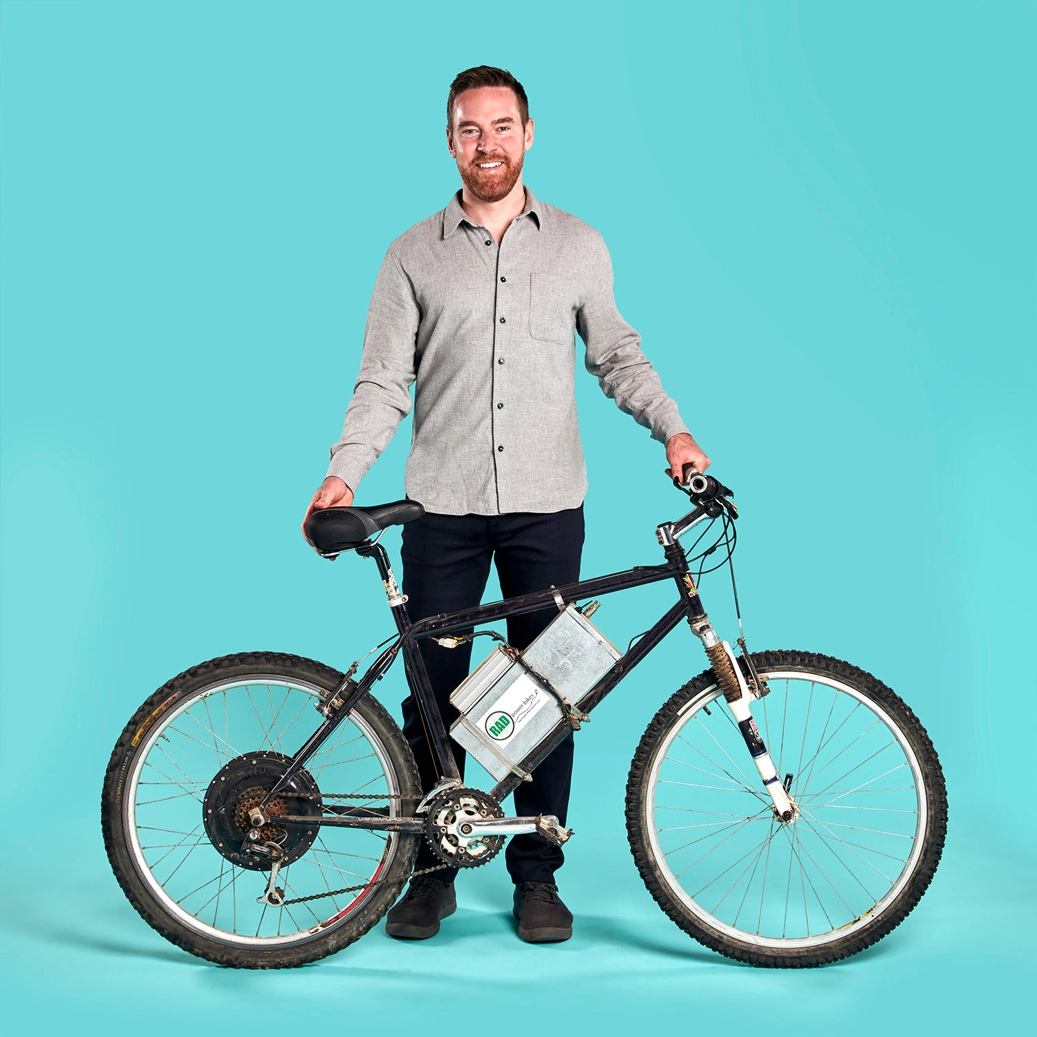 A man stands beside a bicycle fitted with a large hub motor in front of a teal backdrop