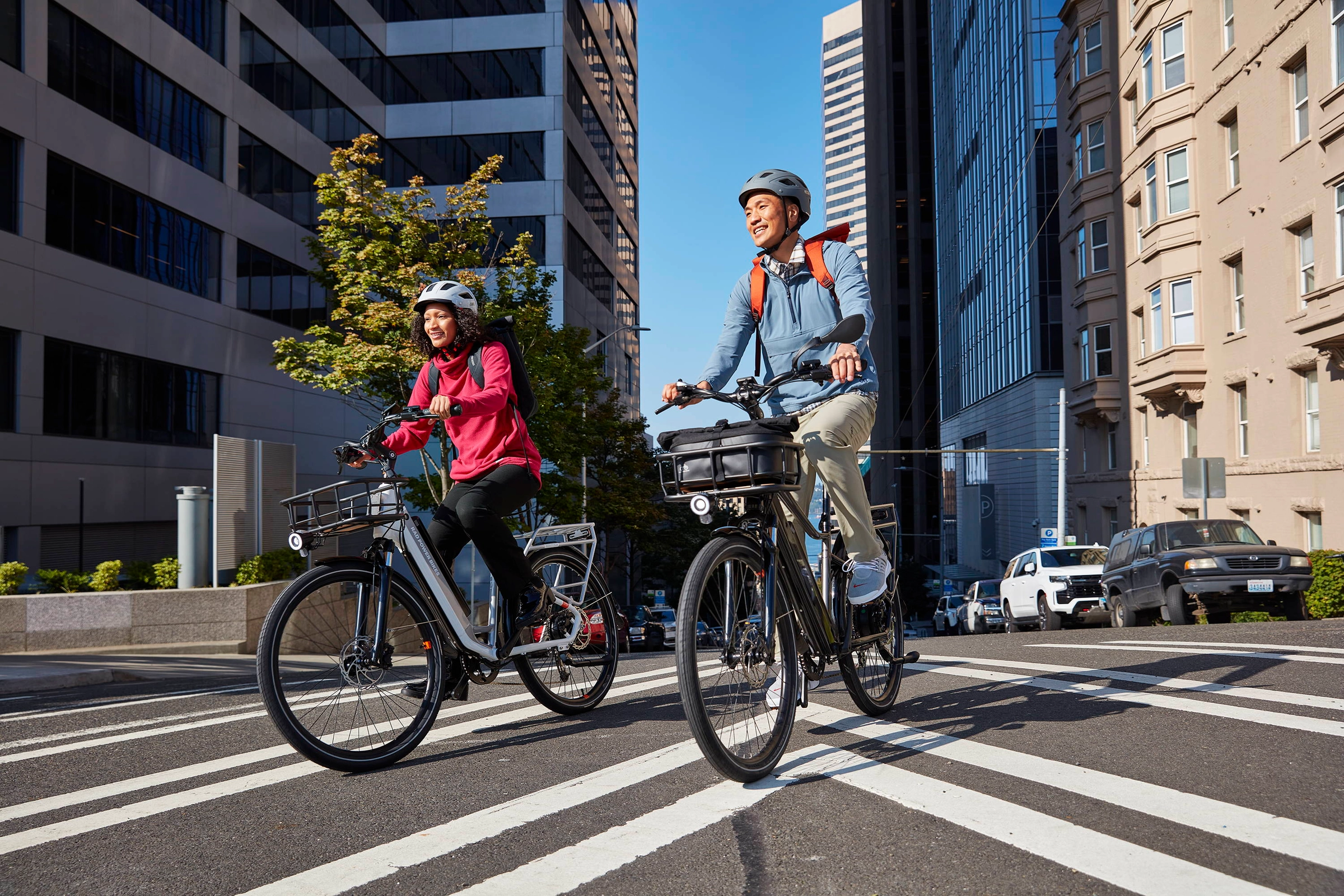 Two riders in bright jackets cross a city street on ebikes between tall buildings